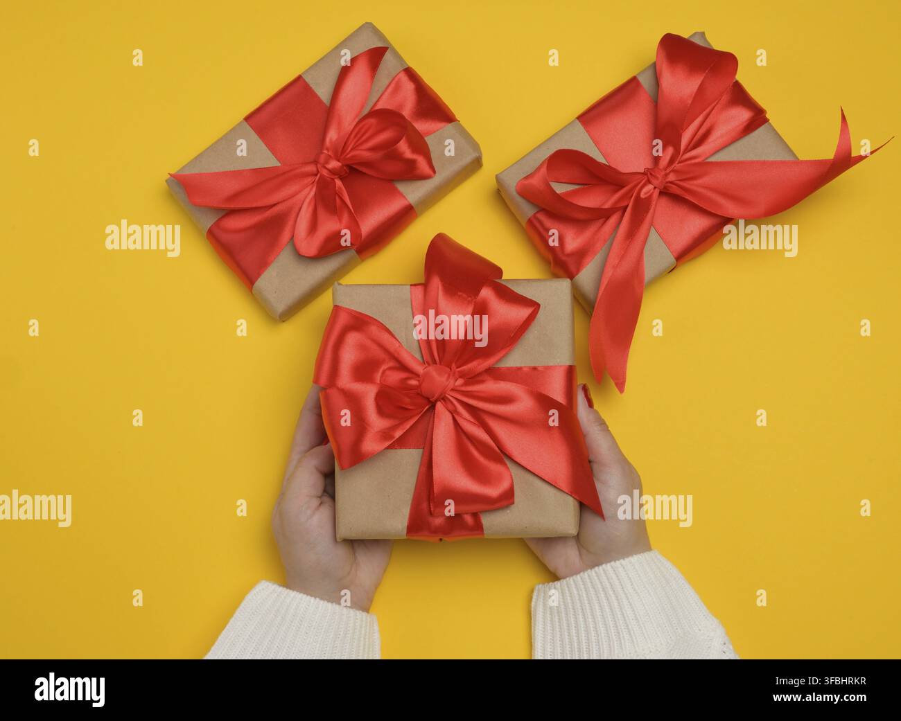 Le mani di una donna in un maglione di lana bianco che tiene un pacco regalo con un nastro di raso rosso Foto Stock