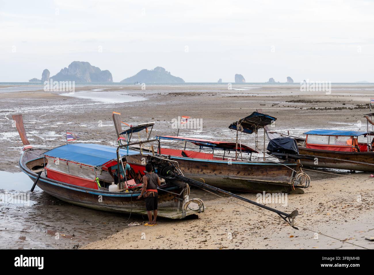 KHLONG MUANG, THAILANDIA – 19 FEBBRAIO 2026: Persone curano barche a lunga carena arenate con la bassa marea, con isole di calcare visibili al largo. Foto Stock