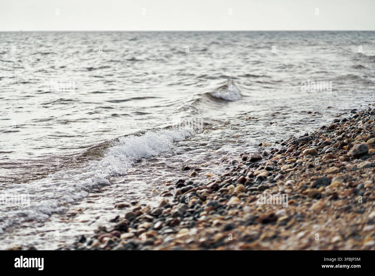 Piccole onde gentili che si infrangono su una riva ghiaiosa colorata con acqua limpida che increspa su pietre lisce lungo la costa della Grecia. Foto Stock
