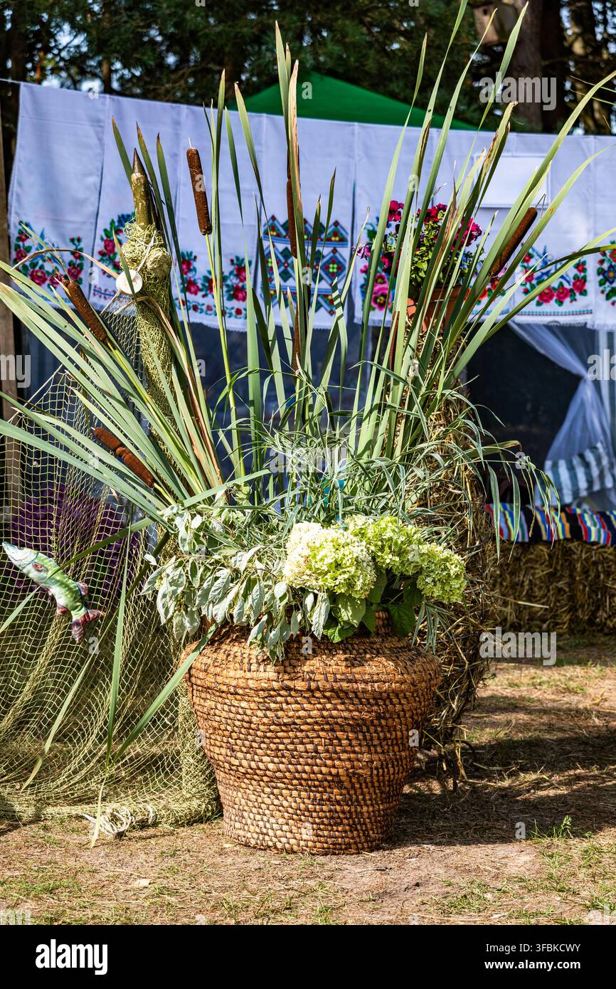 Ampio cesto in vimini intrecciato a mano pieno di canne verdi, tife e fiori freschi, posizionato all'aperto su uno sfondo di tessuti ricamati Foto Stock