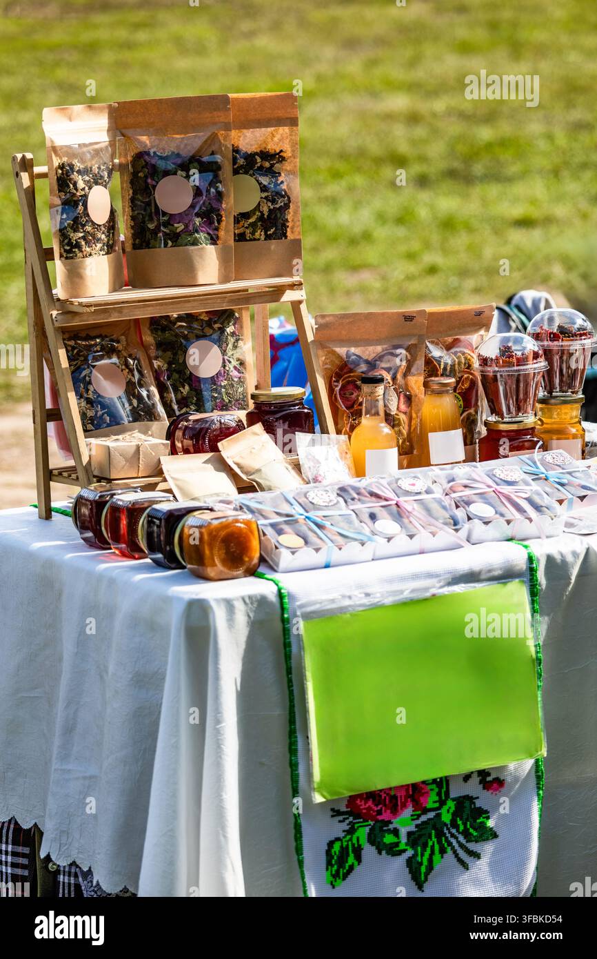 Esposizione di bustine di tè alle erbe biologiche in imballaggi artigianali, barattoli di miele e marmellata naturale disposti su una tovaglia bianca in un evento di mercato contadino all'aperto. Foto Stock