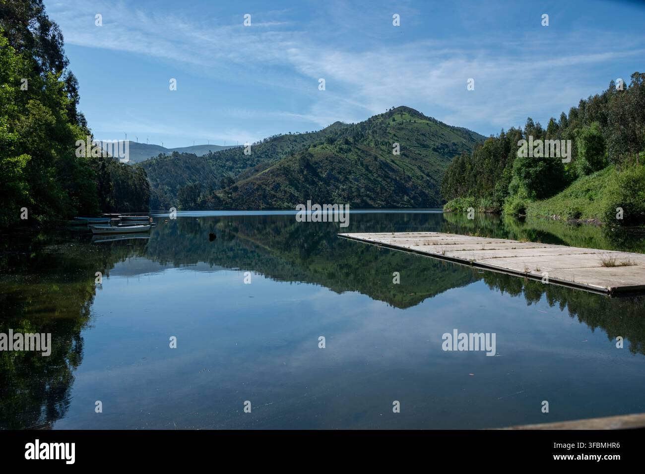 Lago calmo riflette colline lontane e cielo. Gli alberi incorniciano le acque immobili, esaltando la serenità. Nuvole blu fluttuano sopra le cime verdi. La piattaforma di legno riposa dolcemente Foto Stock