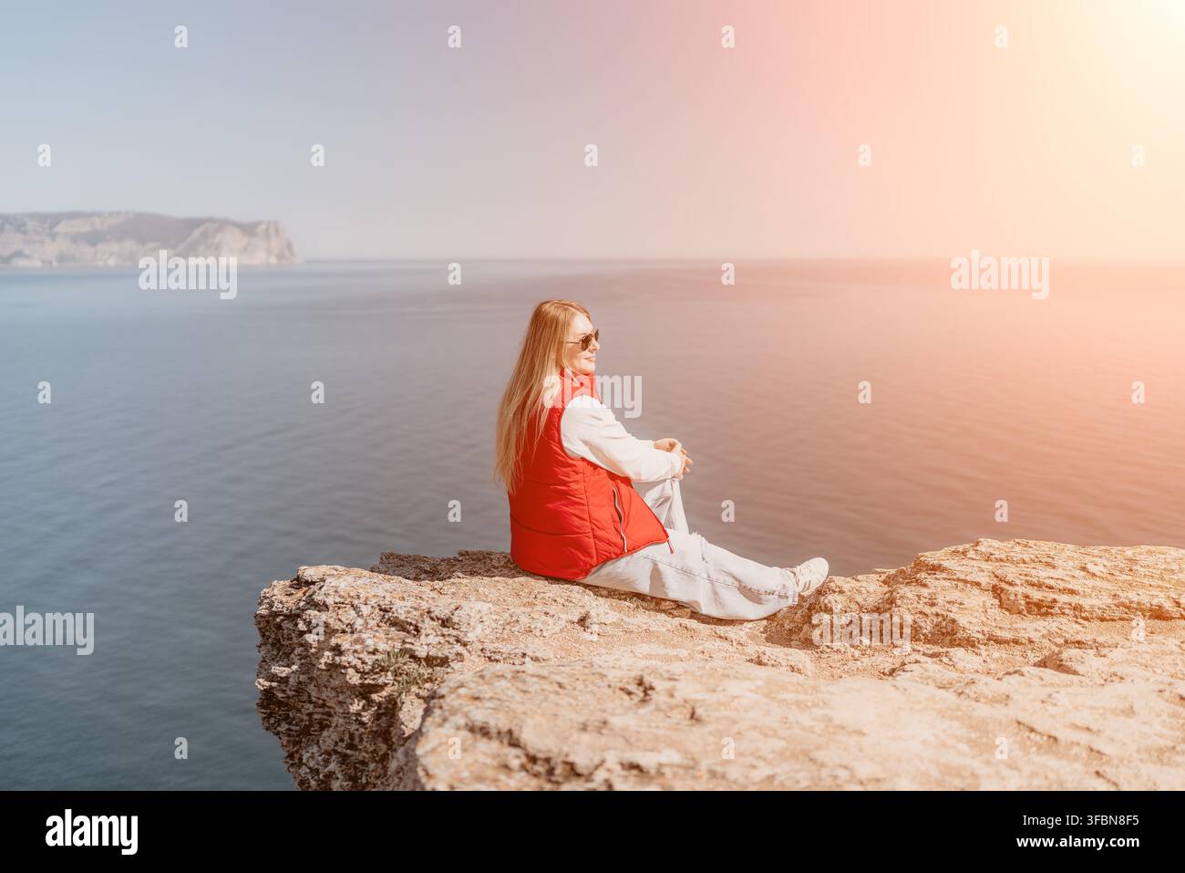 Donna seduta su un bordo roccioso di una scogliera sull'oceano, indossa un giubbotto rosso, si affaccia sull'acqua blu calma con spazio libero Foto Stock