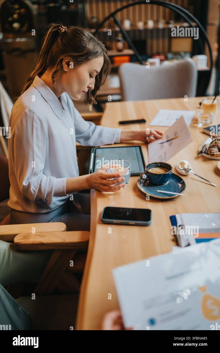 Donna d'affari che esamina documenti e un tablet mentre beve caffè durante un incontro informale in un bar Foto Stock