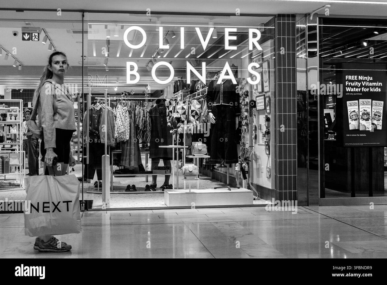Londra UK, 24 agosto 2026, Scatto in bianco e nero di una giovane donna in piedi davanti alla vetrina di un negozio Oliver Bonas con una borsa di Next in mano Foto Stock