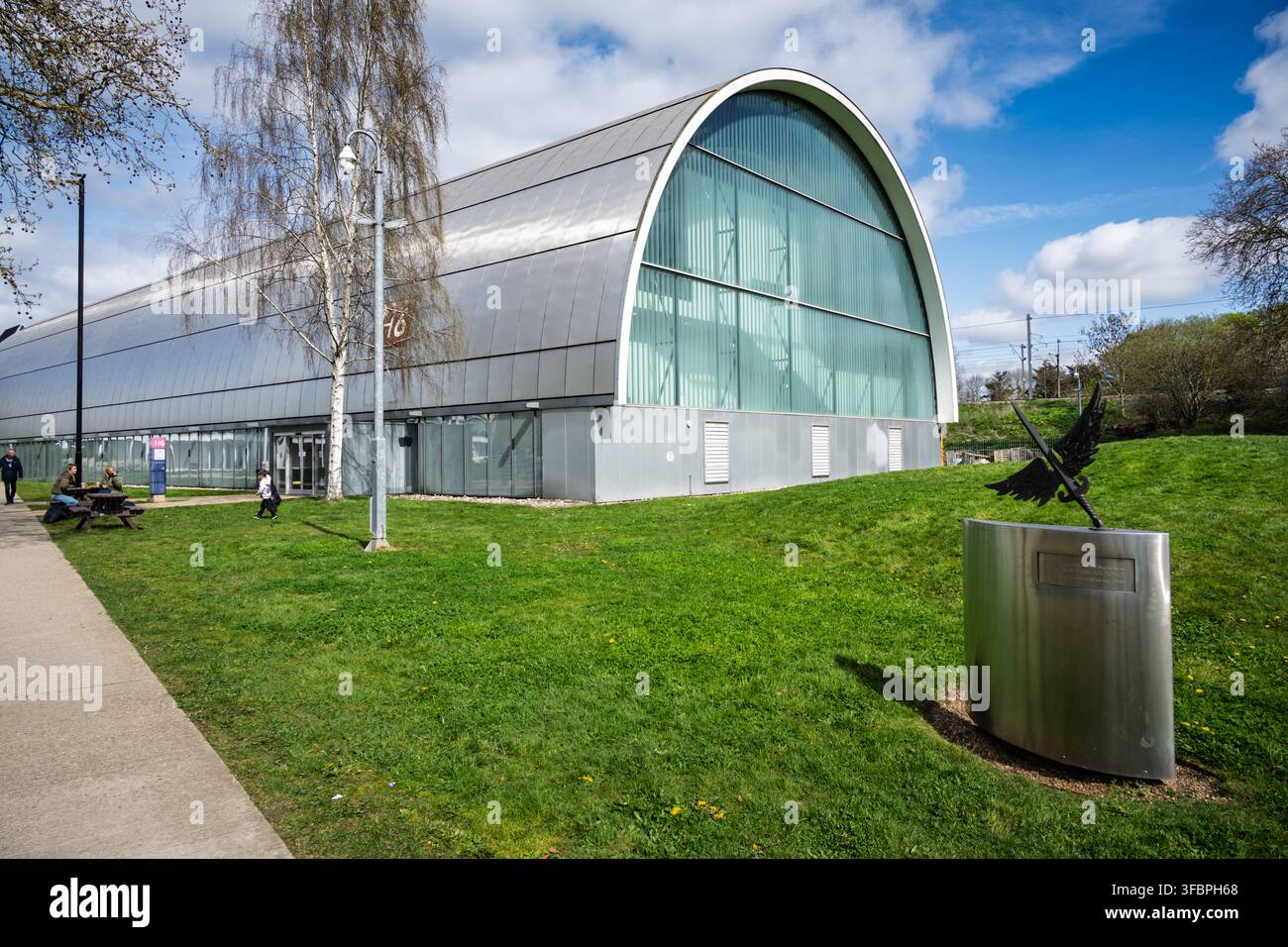 L'esterno ad arco moderno dell'Hangar 6 accanto alla scultura monumentale a forma di gru metallica sul terreno del Royal Air Force Museum di Londra Foto Stock
