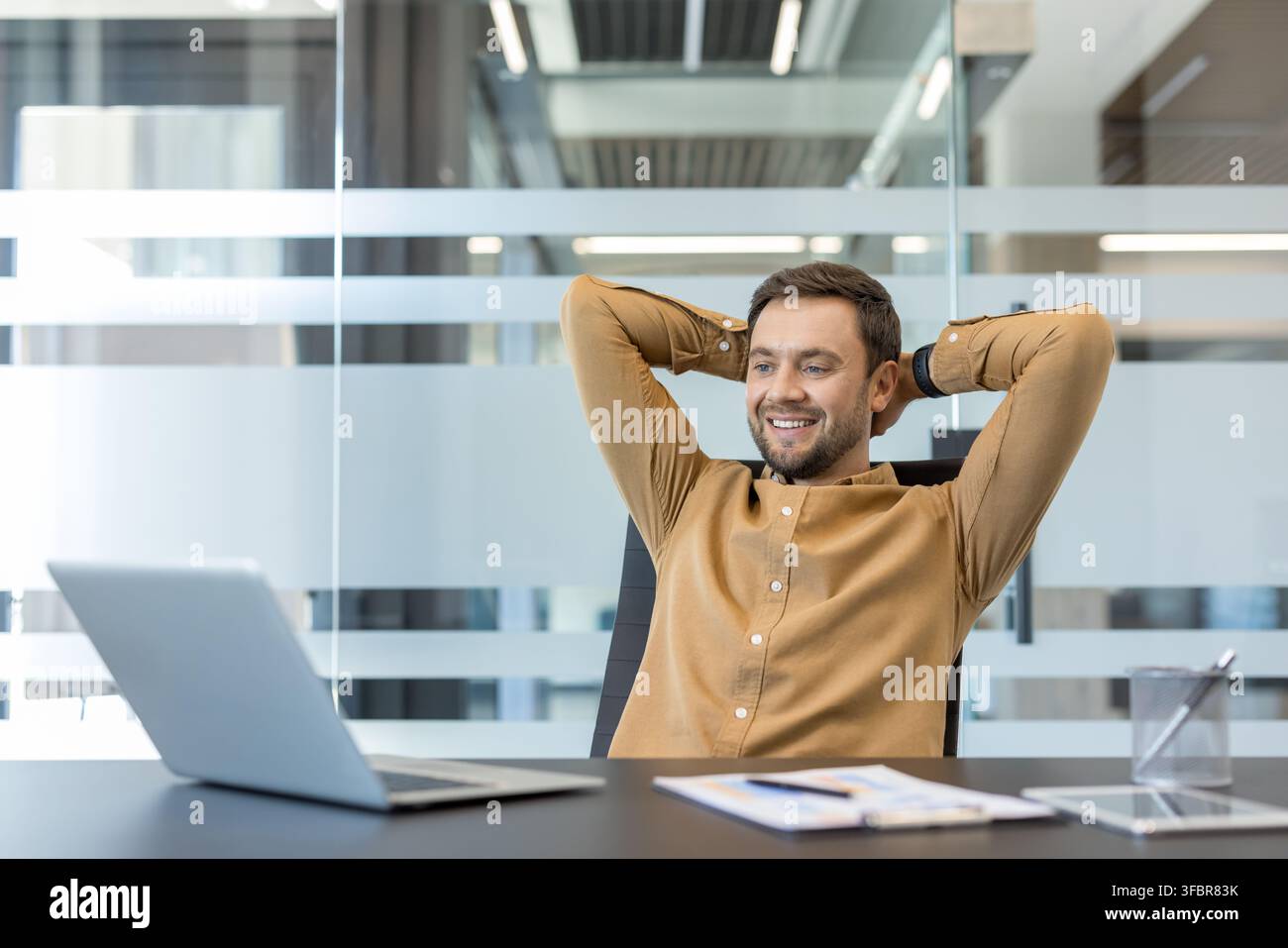Uomo d'affari sorridente appoggiato allo schienale della sua sedia da ufficio con le mani dietro la testa, godendosi un momento di relax e soddisfazione dopo aver raggiunto obiettivi e completato compiti Foto Stock