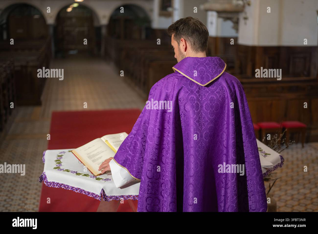 Un prete si trova in una chiesa e legge da una bibbia. La chiesa ha panche di legno e un tappeto rosso. Foto Stock