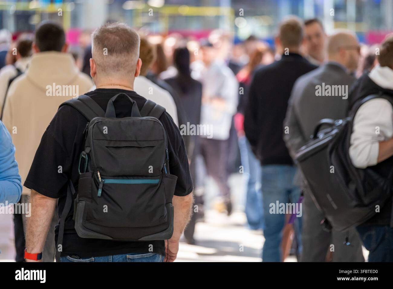 Uomo con uno zaino nero attraversa una folla indaffarata di un'esposizione al coperto sotto una luce moderna e luminosa. Foto Stock