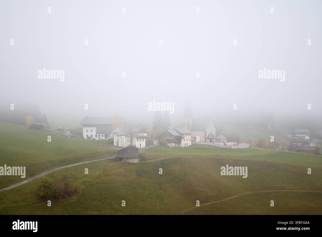 La chiesa di Santa Maddalena emerge dalla fitta nebbia mattutina nella Val di Funes, Alto Adige. Paesaggio alpino atmosferico con fattorie tradizionali e prati nebbiosi Foto Stock