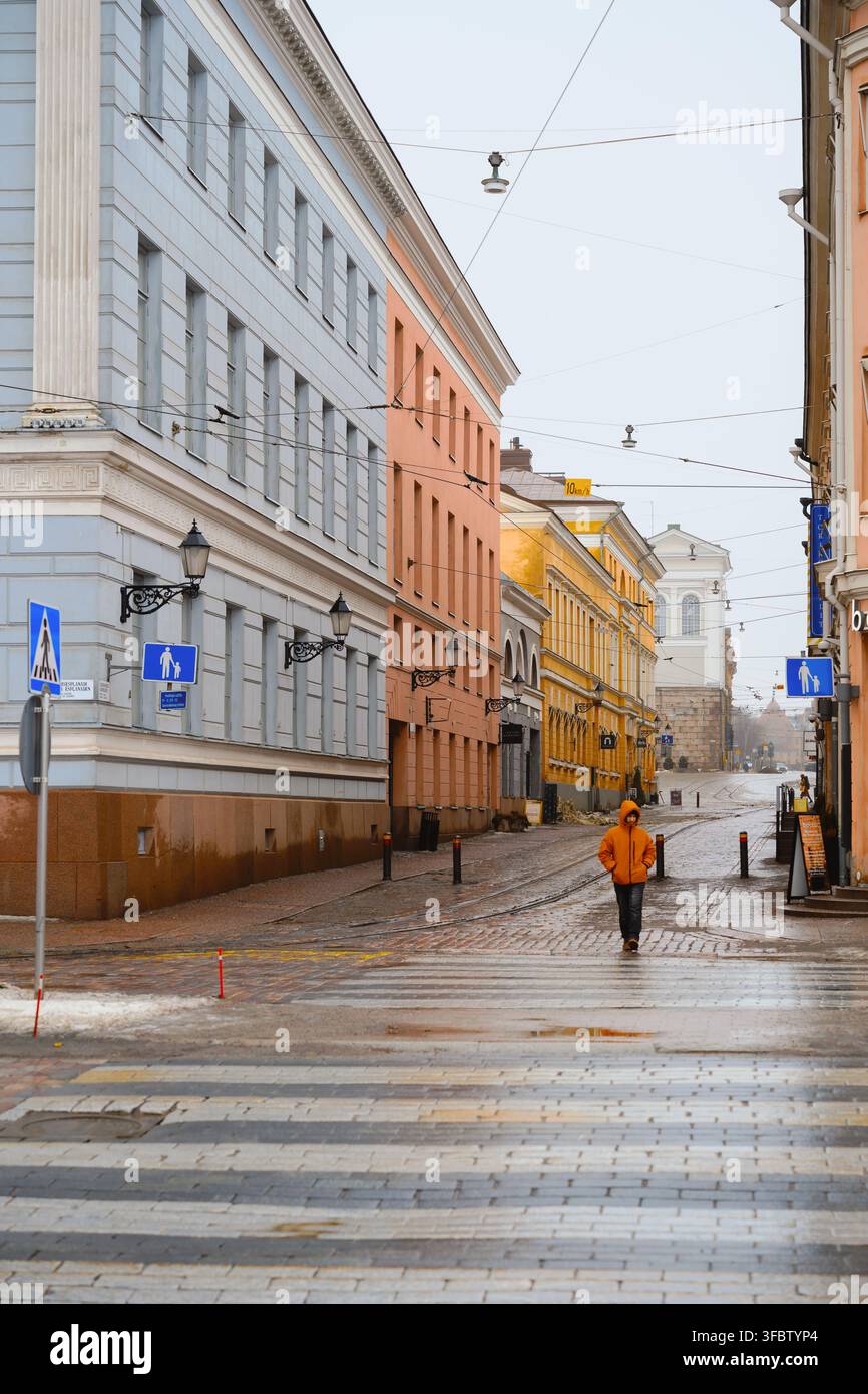 Helsinki, Finlandia - 1° marzo 2026: Pedone con giacca arancione cammina lungo una strada bagnata fiancheggiata da edifici storici. Foto Stock