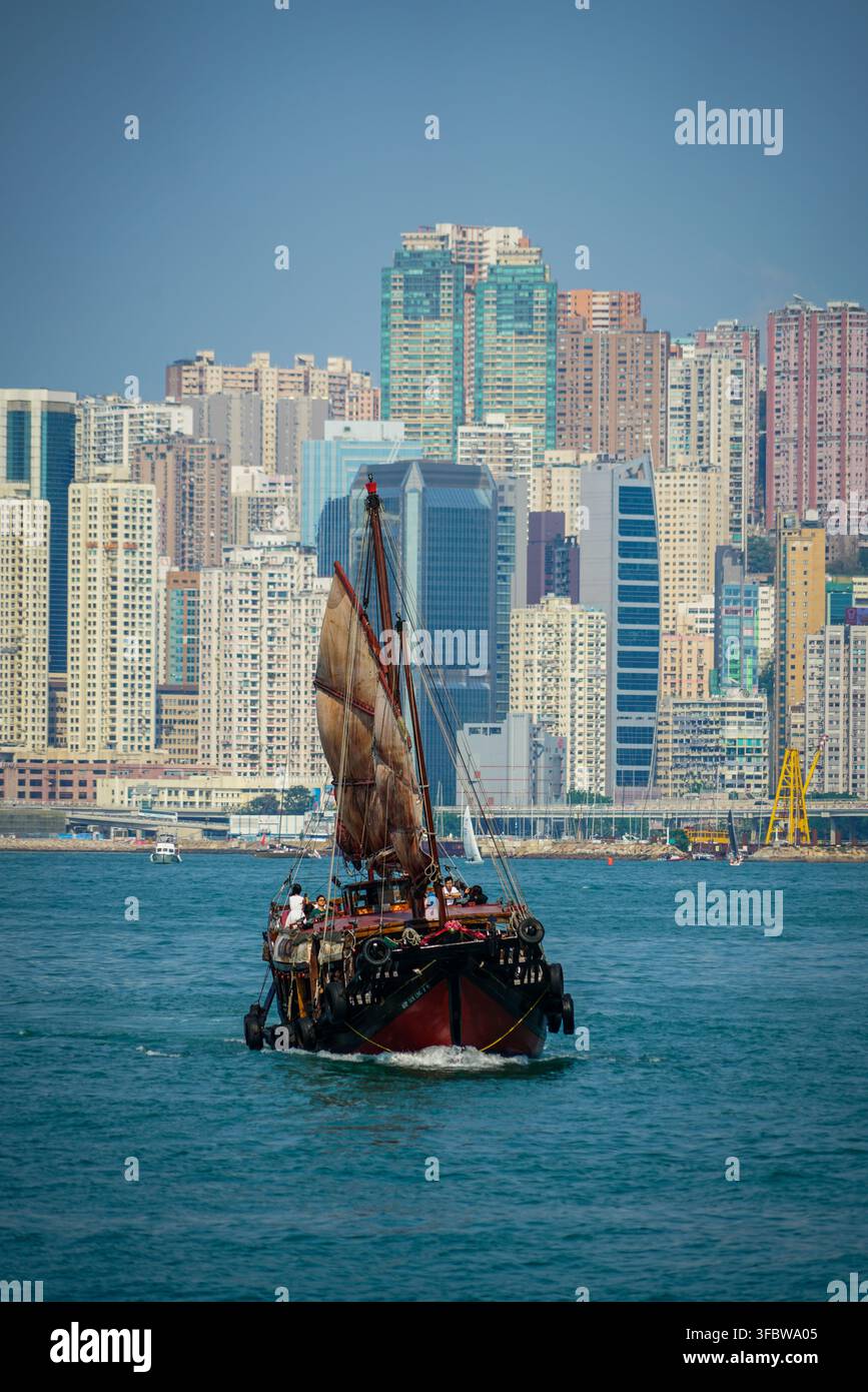 Un tradizionale barca junk rossa e nera con una grande vela marrone arrotolata scivola sulle acque blu profonde del Victoria Harbour. In lontananza, il d Foto Stock