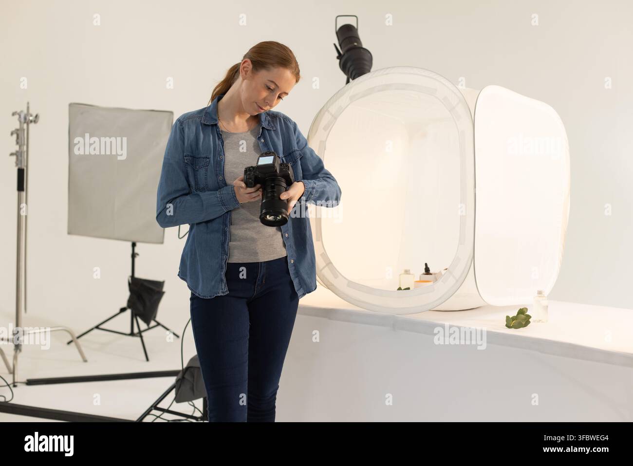 Donna sulla ventina che controlla le impostazioni della DSLR mentre si appoggia a un tavolo vicino a una tenda luminosa con bottiglie Foto Stock