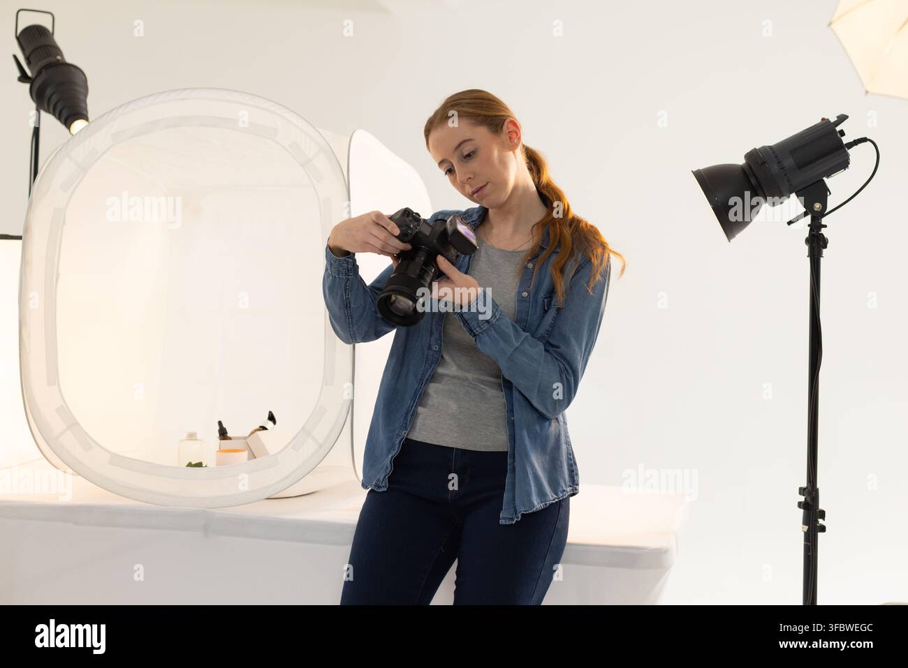 Fotografa adulta che ispeziona i controlli della fotocamera DSLR mentre si appoggia su un tavolo vicino a una tenda luminosa Foto Stock