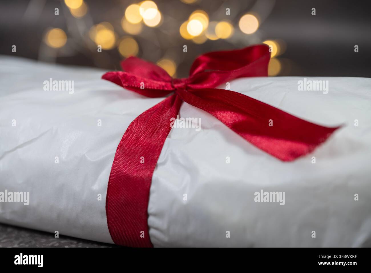 Un primo piano di un lungo pacco avvolto in carta bianca e legato con un fiocco di raso rosso festoso su uno sfondo scuro con luci sfocate a bokeh. Foto Stock