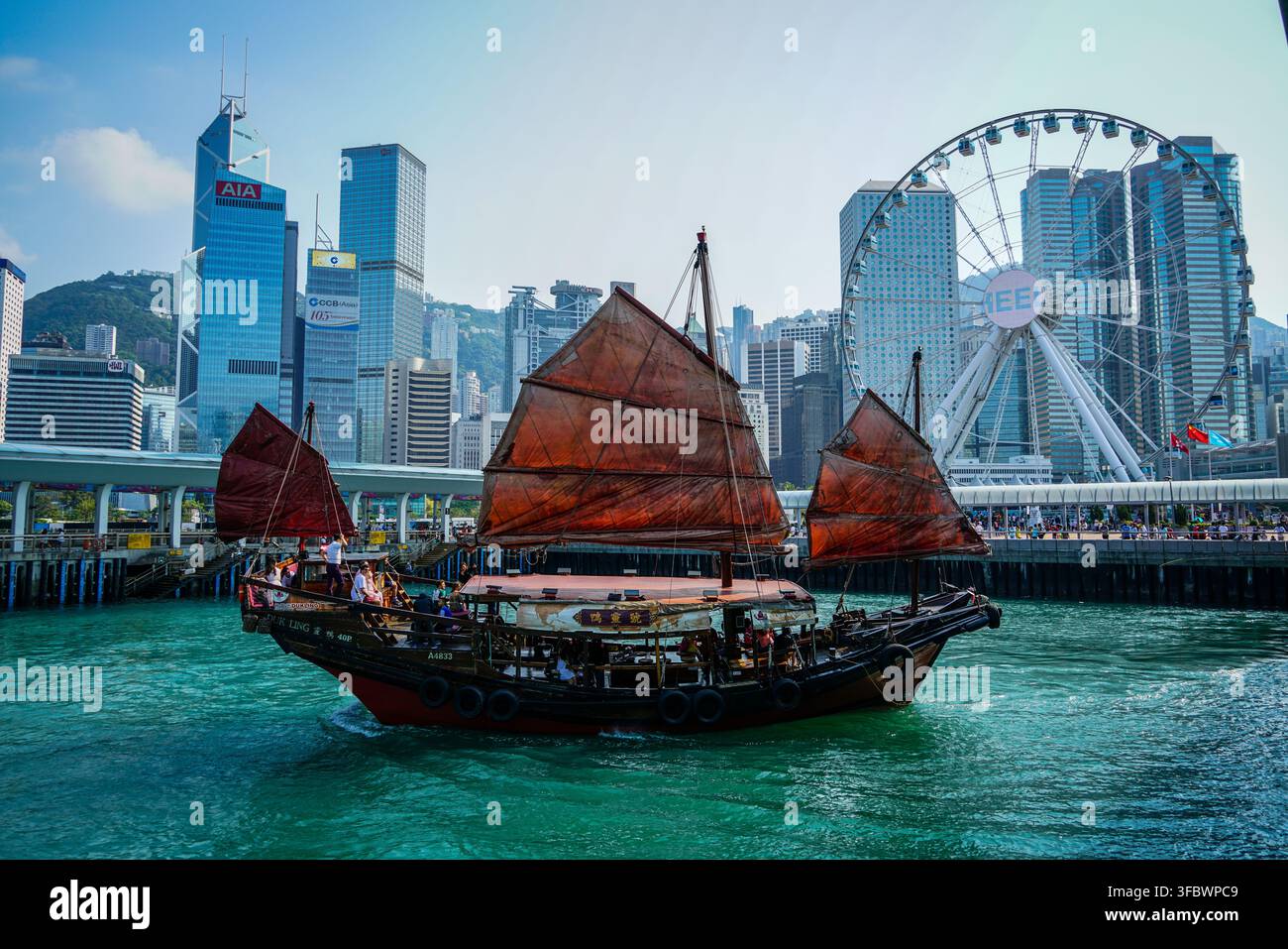 Una giunca Aqua Luna con vela rossa si muove sull'acqua turchese del Victoria Harbour, con la ruota panoramica di Hong Kong e diversi grattacieli, i Foto Stock