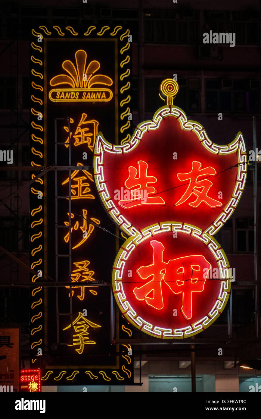 Un'insegna al neon verticale nera e gialla per "Sands Sauna" è appesa accanto a un cartello rosso a forma di scudo e a un cartello rosso circolare, entrambi con caratteri cinesi a Foto Stock