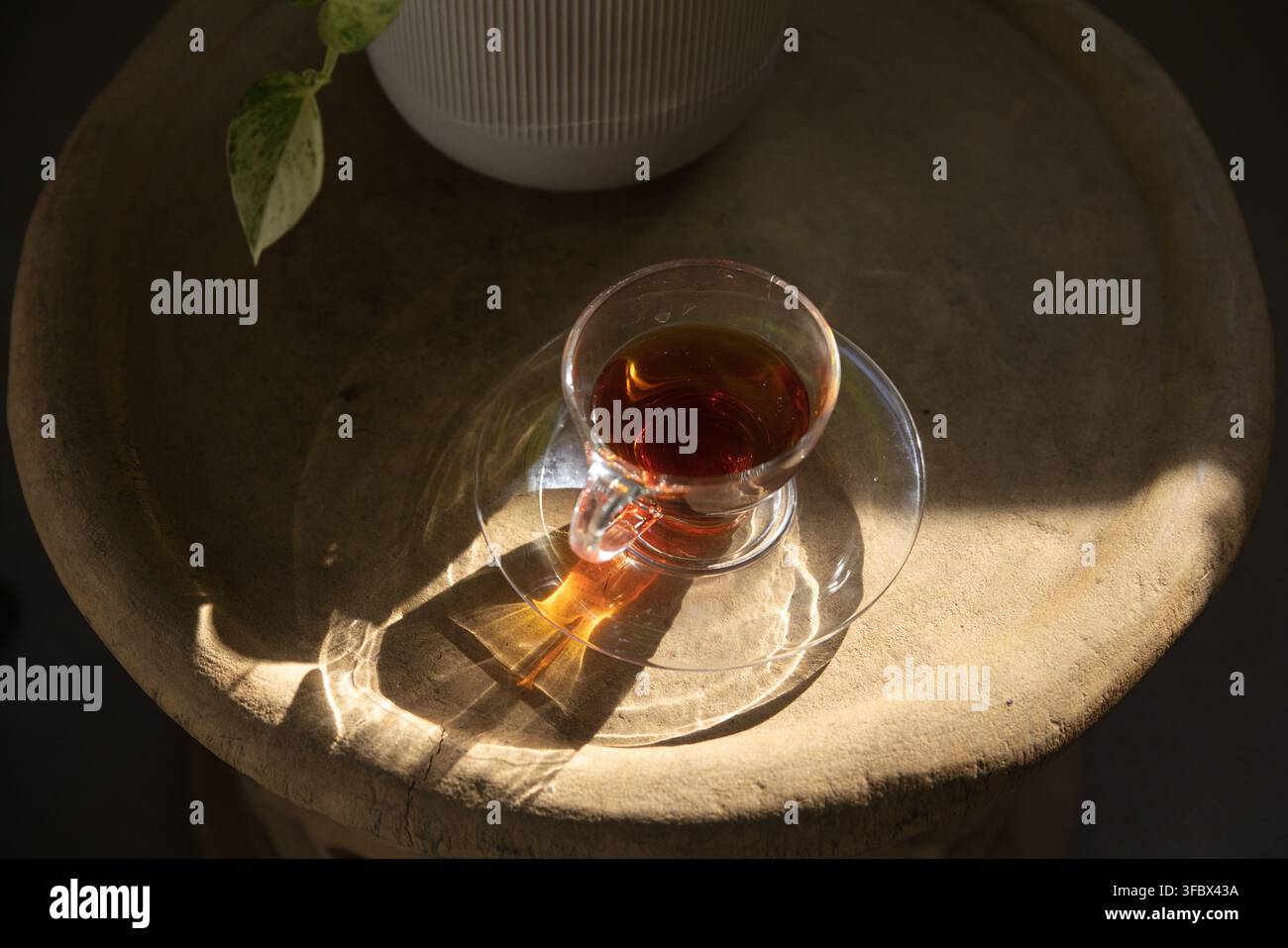 Tazza da tè in vetro trasparente su piattino è posata su un tavolo ruvido mentre la luce del sole proietta motivi rifratti Foto Stock