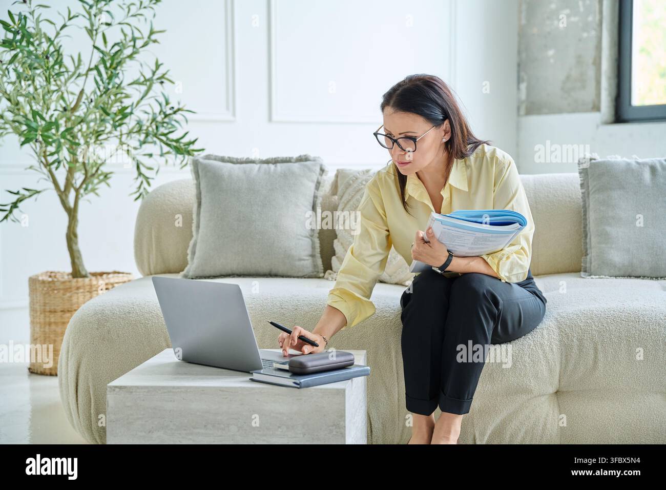 Psicologa matura che tiene una cartella e usa un laptop sul divano a casa Foto Stock