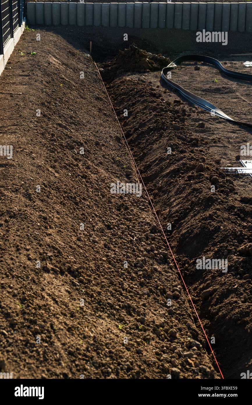 Scavatura di una trincea per l'installazione di un sistema di drenaggio in un cantiere edile residenziale. L'area mostra terra appena scavata con una chiara demarcazione, che indica la preparazione per i lavori di idraulica. Foto Stock