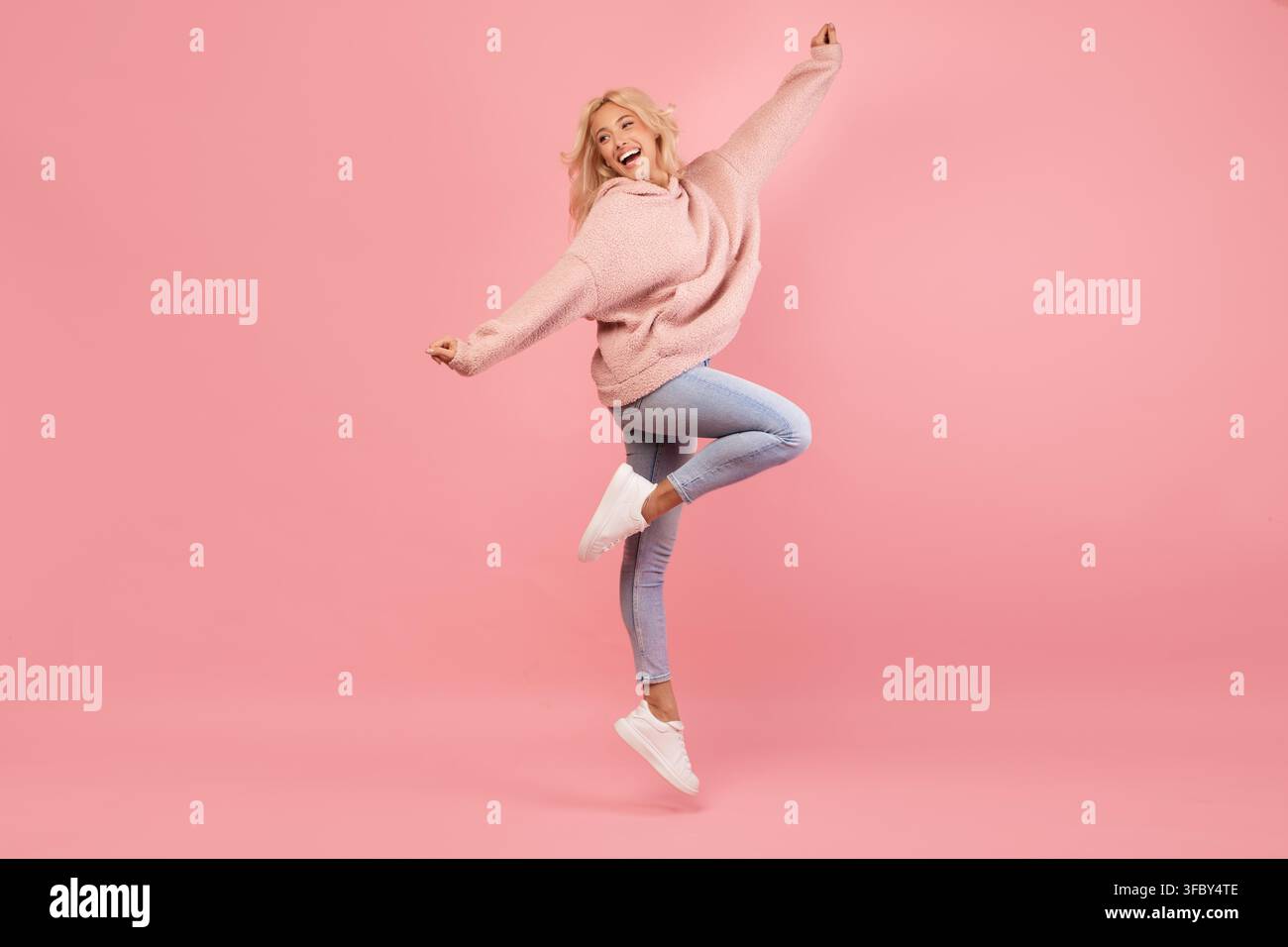 Giovane donna caucasica eccitata che salta gioiosamente in una felpa con cappuccio elegante, sfondo rosa Foto Stock