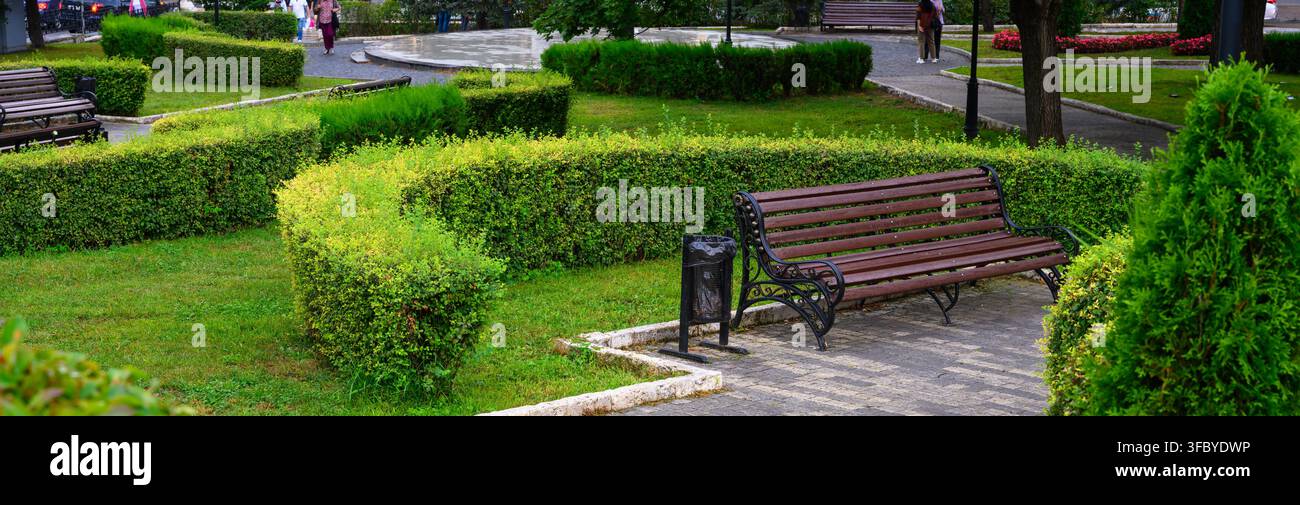 Panchina di legno vuota in un parco pubblico verde a Pyatigorsk, Russia. Paesaggio urbano con siepi curate, erba e vialetto lastricato. Infrastruttura da giardino cittadino Foto Stock