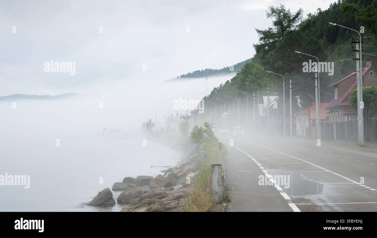 Nebbia fitta su una strada costiera con auto in movimento al Lago Baikal, Russia. Condizioni meteorologiche a bassa visibilità su una strada lakeside con case e strade Foto Stock