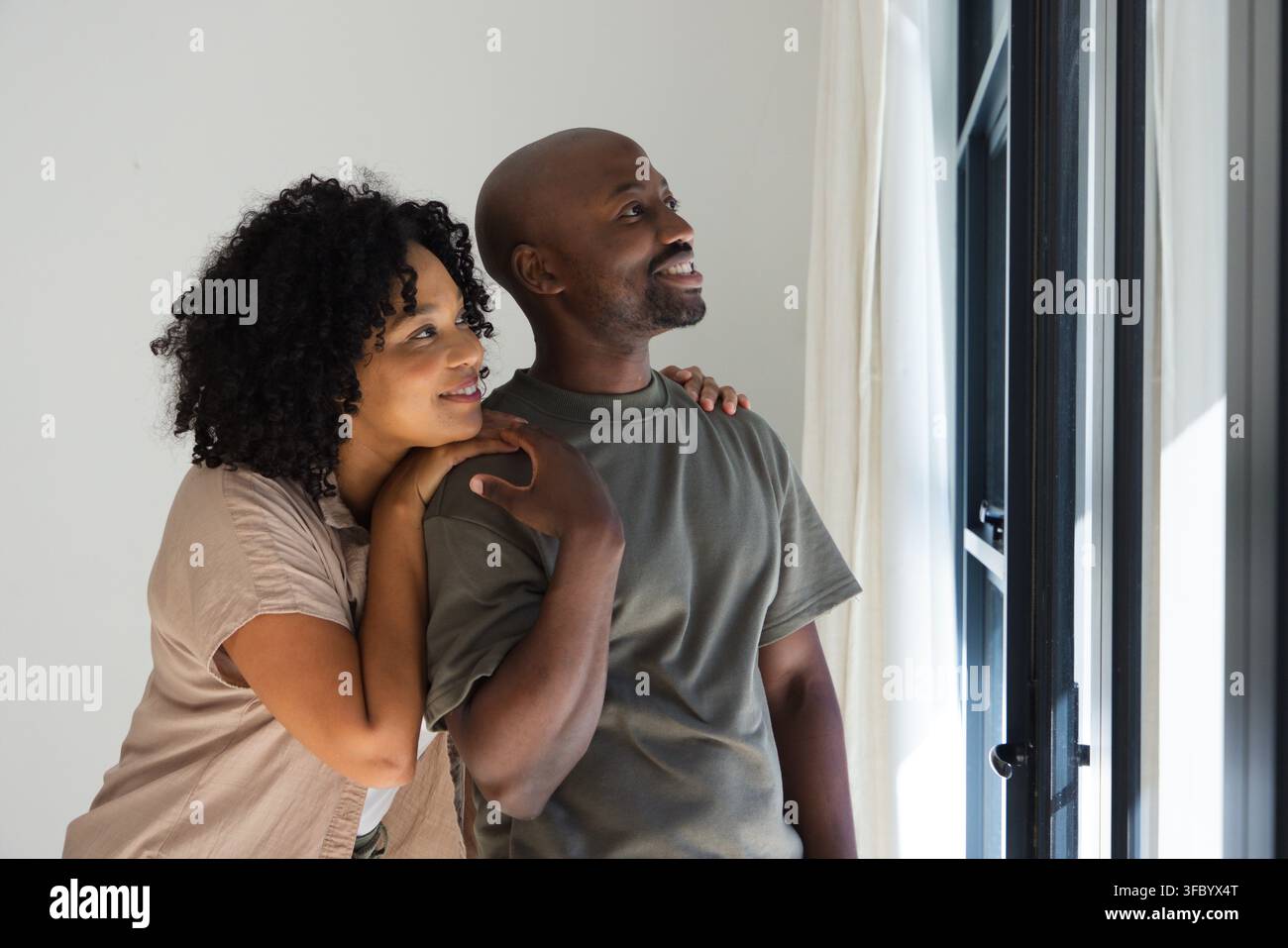 Coppia africana in piedi vicino a una finestra nera con tende bianche, appoggiata sulla spalla in top oliva marrone chiaro Foto Stock