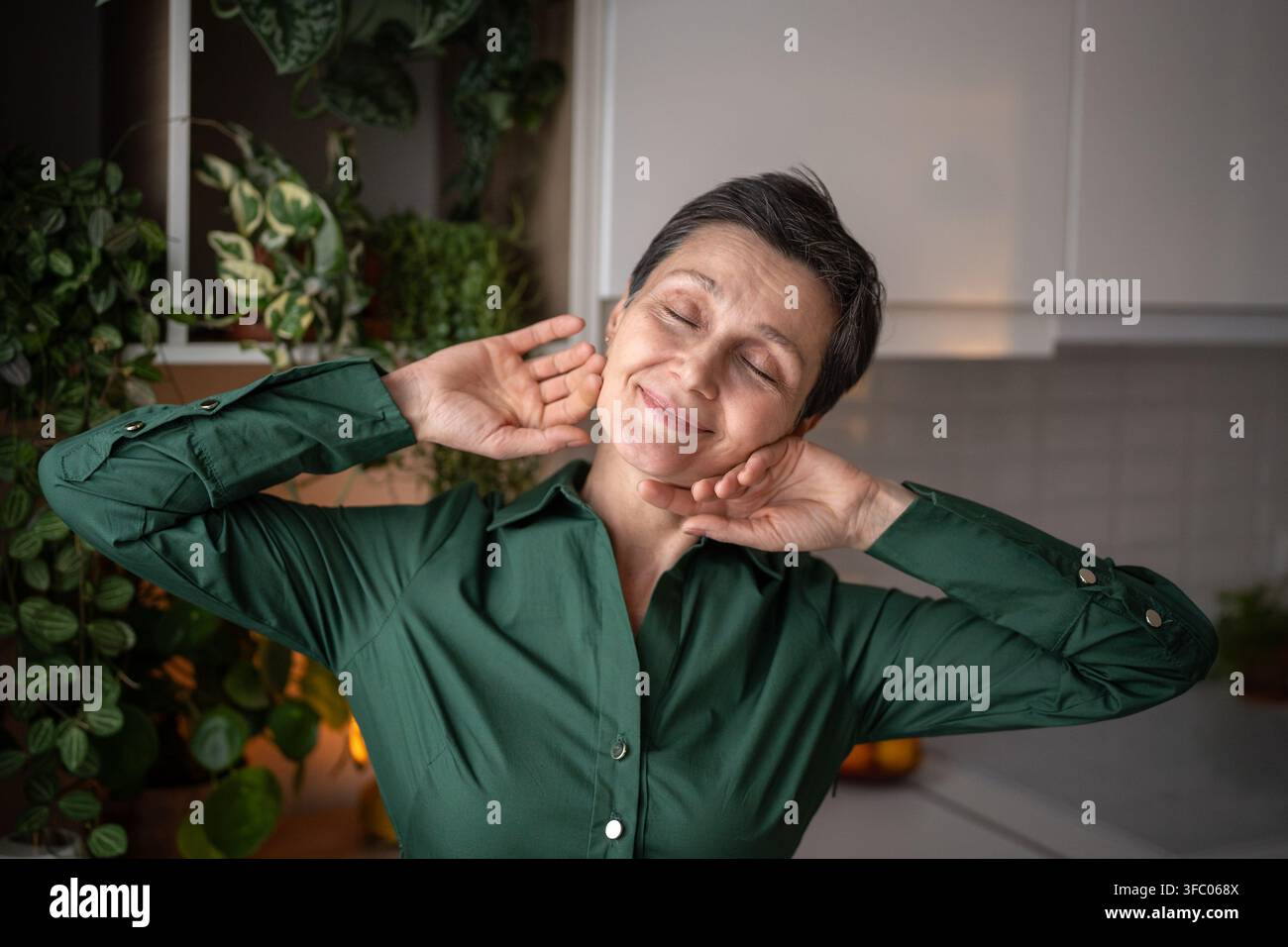 Una donna matura felice e soddisfatta che si gode la luce del sole mattutino nell'accogliente cucina casalinga durante i fine settimana Foto Stock