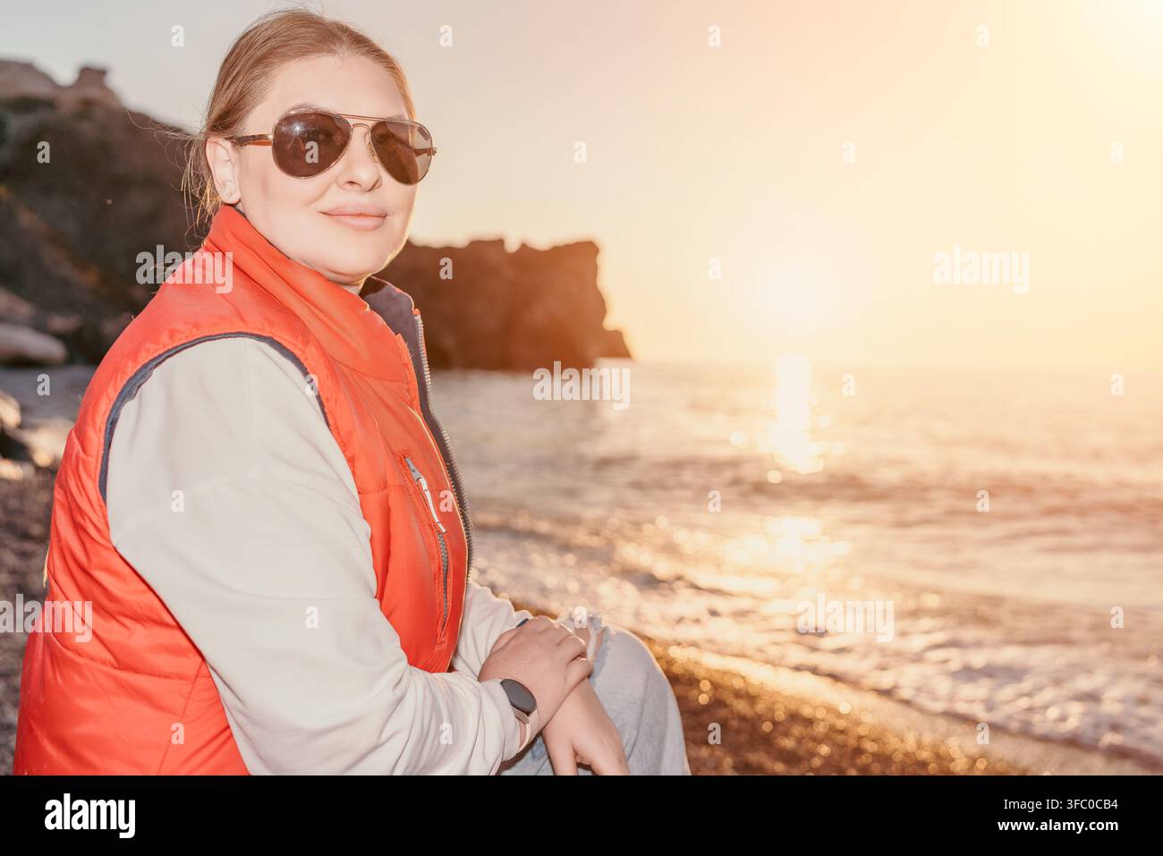 Donna spiaggia tramonto donna sorridente con occhiali da sole e gilet rosso seduta sulla riva di ciottoli vicino all'oceano nell'ora dorata con spazio di copia Foto Stock