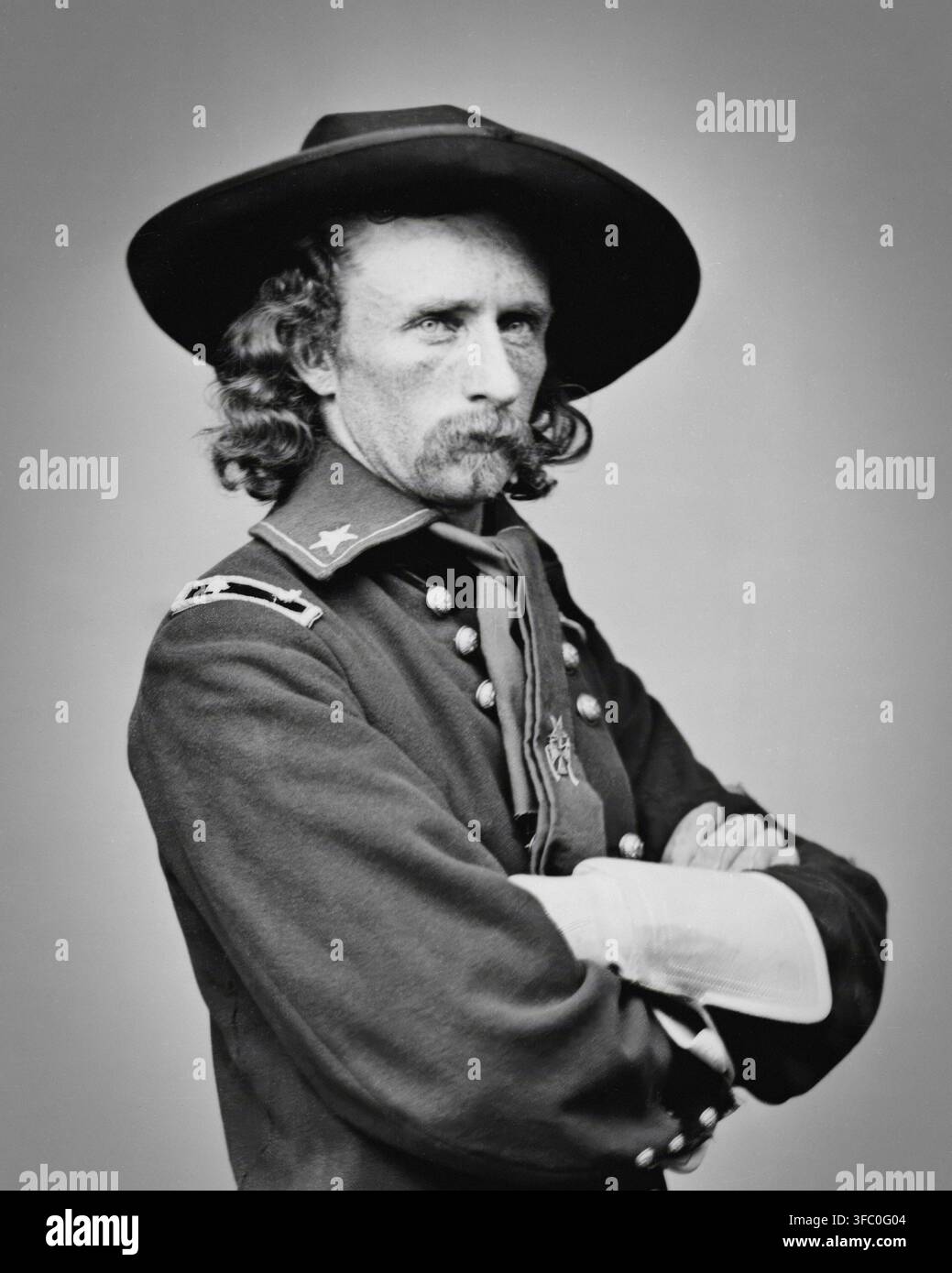 Un ritratto in bianco e nero di George Armstrong Custer, un maggiore generale brevetto, che indossa una uniforme da campo con le braccia incrociate. Questa fotografia, scattata intorno al 1865, mostra Custer con i suoi lunghi capelli e baffi caratteristici, un cappello a tesa larga e una giacca da uniforme adornata con insegne di grado e medaglie. Foto Stock