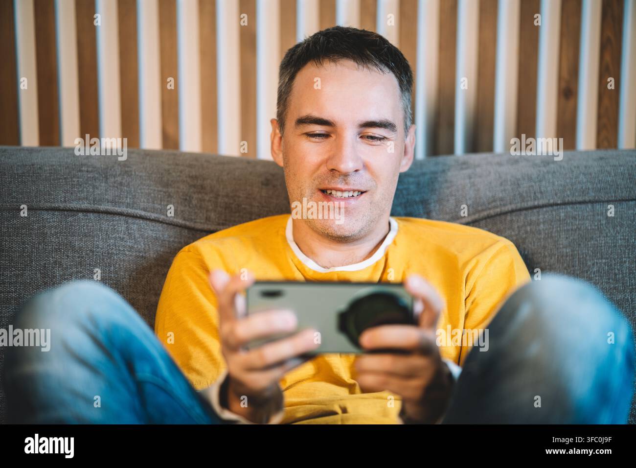 Uomo che gioca a un gioco sullo smartphone seduto sul divano, intrattenimento mobile, scena indoor rilassata. Foto Stock