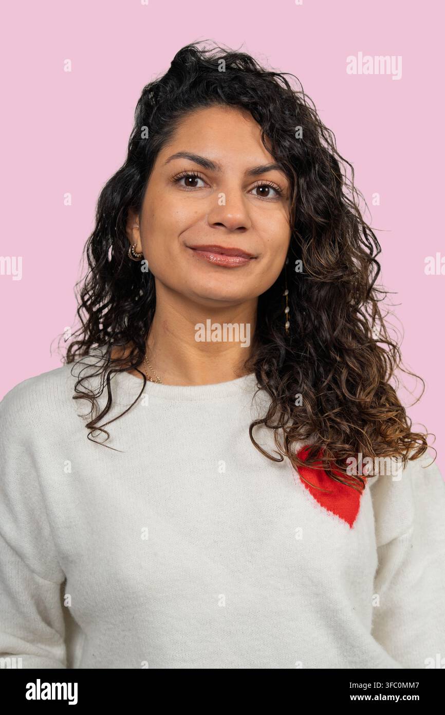 Donna con capelli ricci scuri che posa con un sorriso sottile su uno sfondo rosa solido Foto Stock