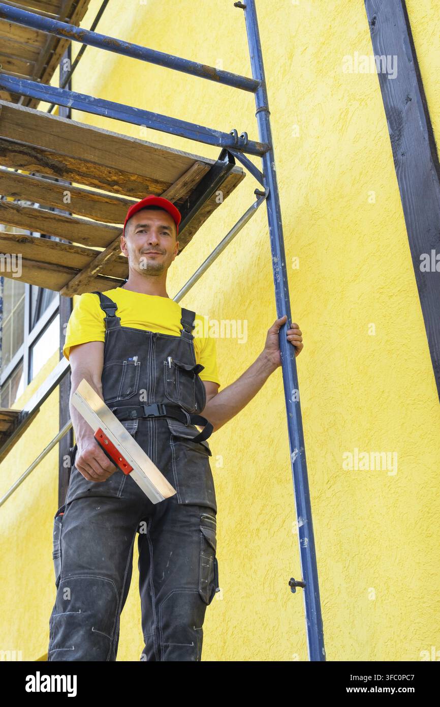 Mastro intonaco con cazzuola su impalcatura, lavori di facciata, applicazione e livellamento di composti, rifiniture esterne, attività di ristrutturazione privata, impianto casa Foto Stock