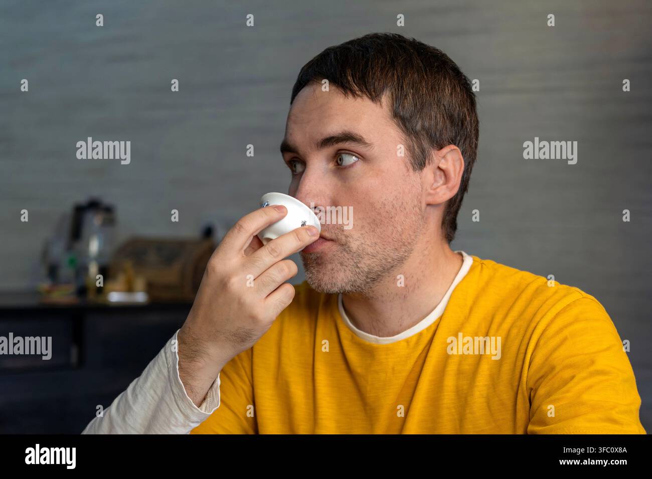 Uomo pensieroso che beve tè da una piccola ciotola o tazza bianca, guardando pensierosamente nel vuoto. Cerimonia del tè, bere consapevole e introspezione silenziosa. Foto Stock