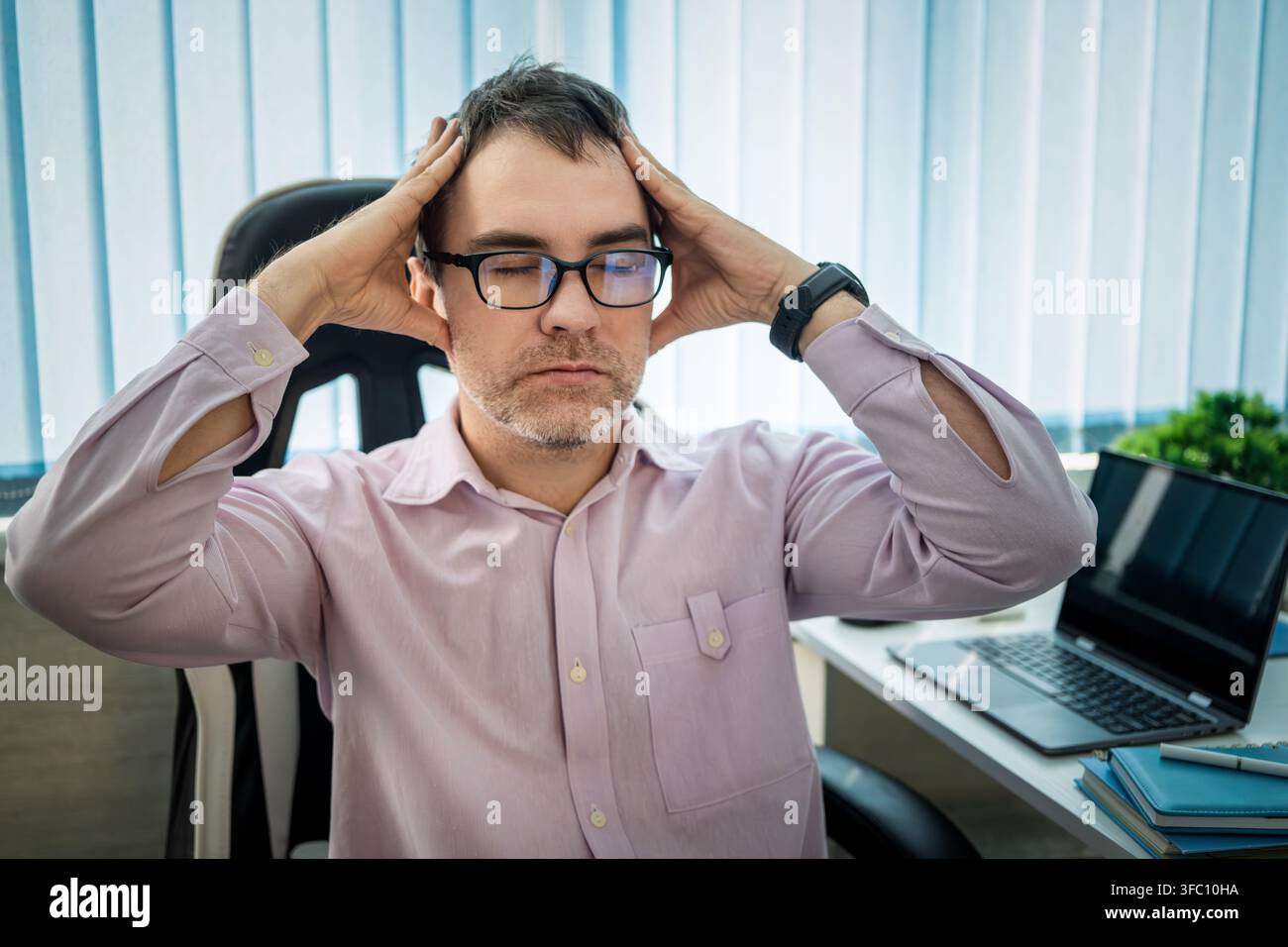 Dipendente d'ufficio stressato che tiene entrambe le mani sulla testa per la frustrazione mentre è seduto alla sua scrivania. Forte mal di testa, esaurimento mentale e pressione sul lavoro Foto Stock