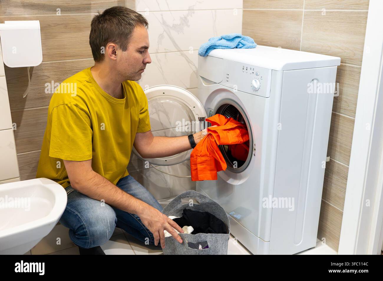 Un uomo con una maglietta gialla carica un indumento arancione in una lavatrice; un asciugamano blu riposa sopra, un cesto della biancheria è nelle vicinanze, in una piastrella compatta Foto Stock