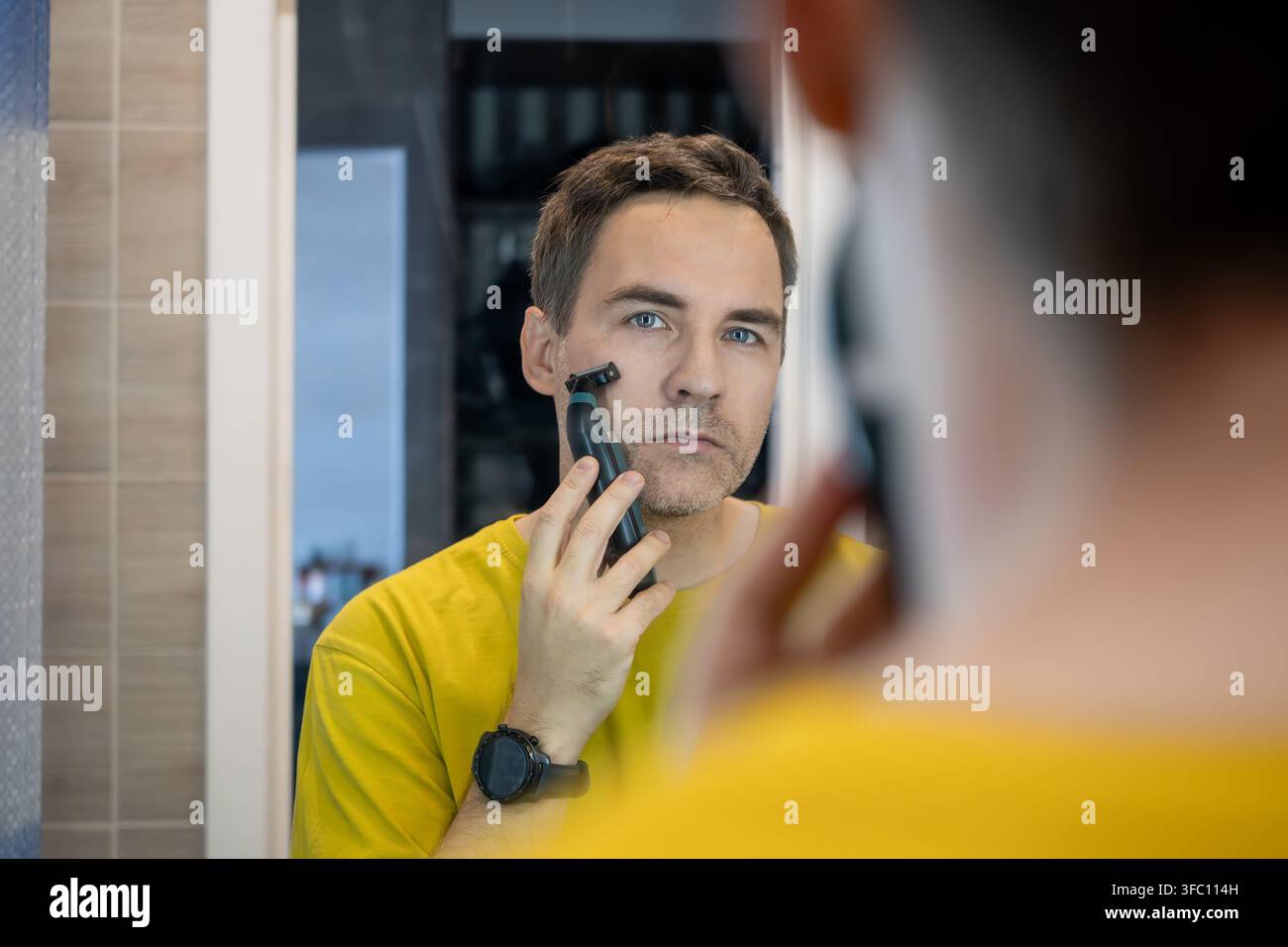 Uomo che si rade con un rasoio elettrico davanti allo specchio. Routine di cura mattutina. Foto Stock