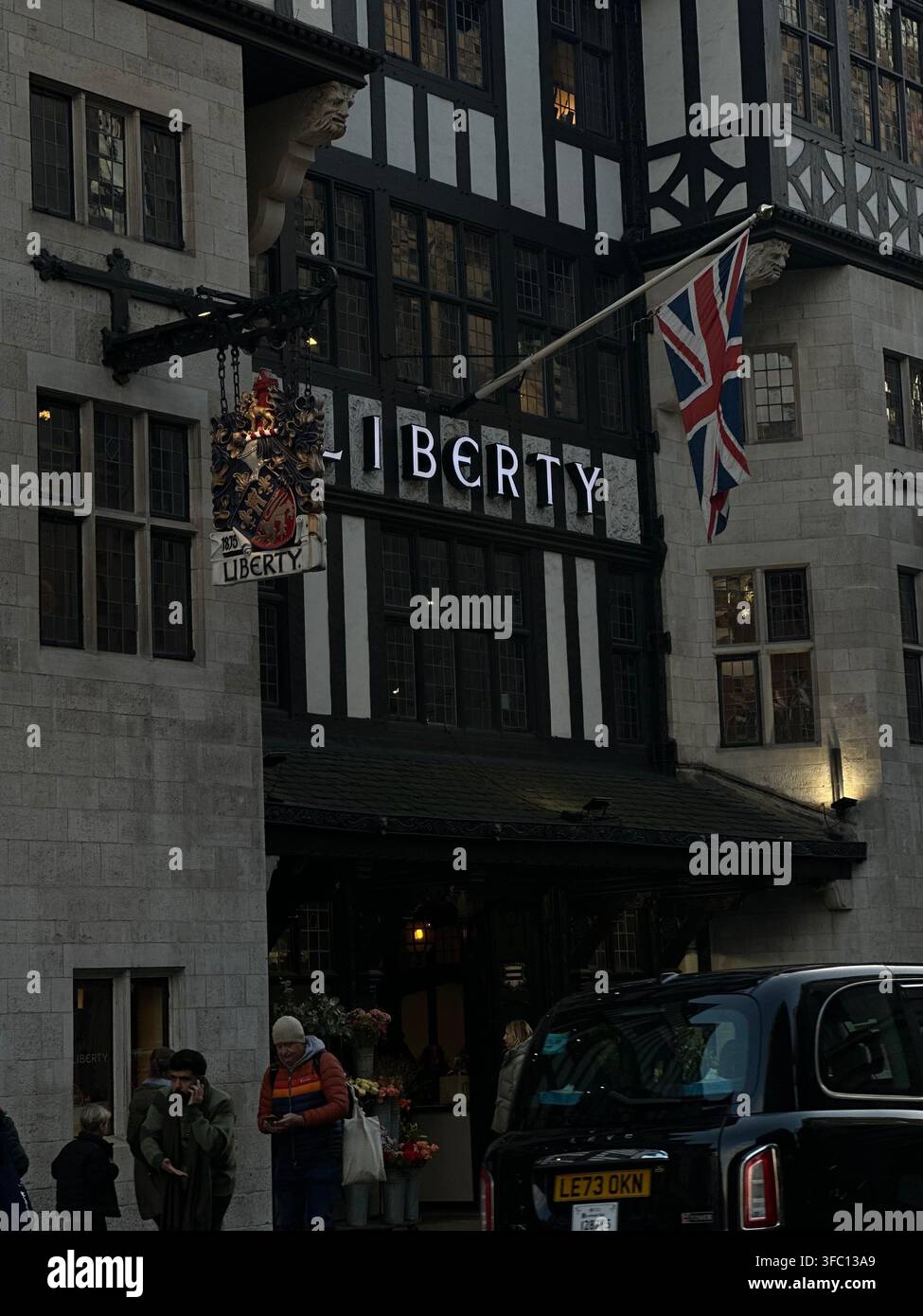 Vista esterna della facciata in stile Tudor Revival del grande magazzino Liberty London con una bandiera Union Jack e un classico taxi nero. Londra, Regno Unito. - Immagine stock catturata con smartphone