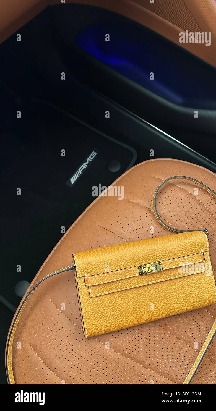Una borsa a tracolla Hermès Kelly To Go in pelle toffee marrone con dettagli dorati esposta su un sedile per auto Mercedes AMG che mostra il logo del marchio - Immagine stock catturata con smartphone