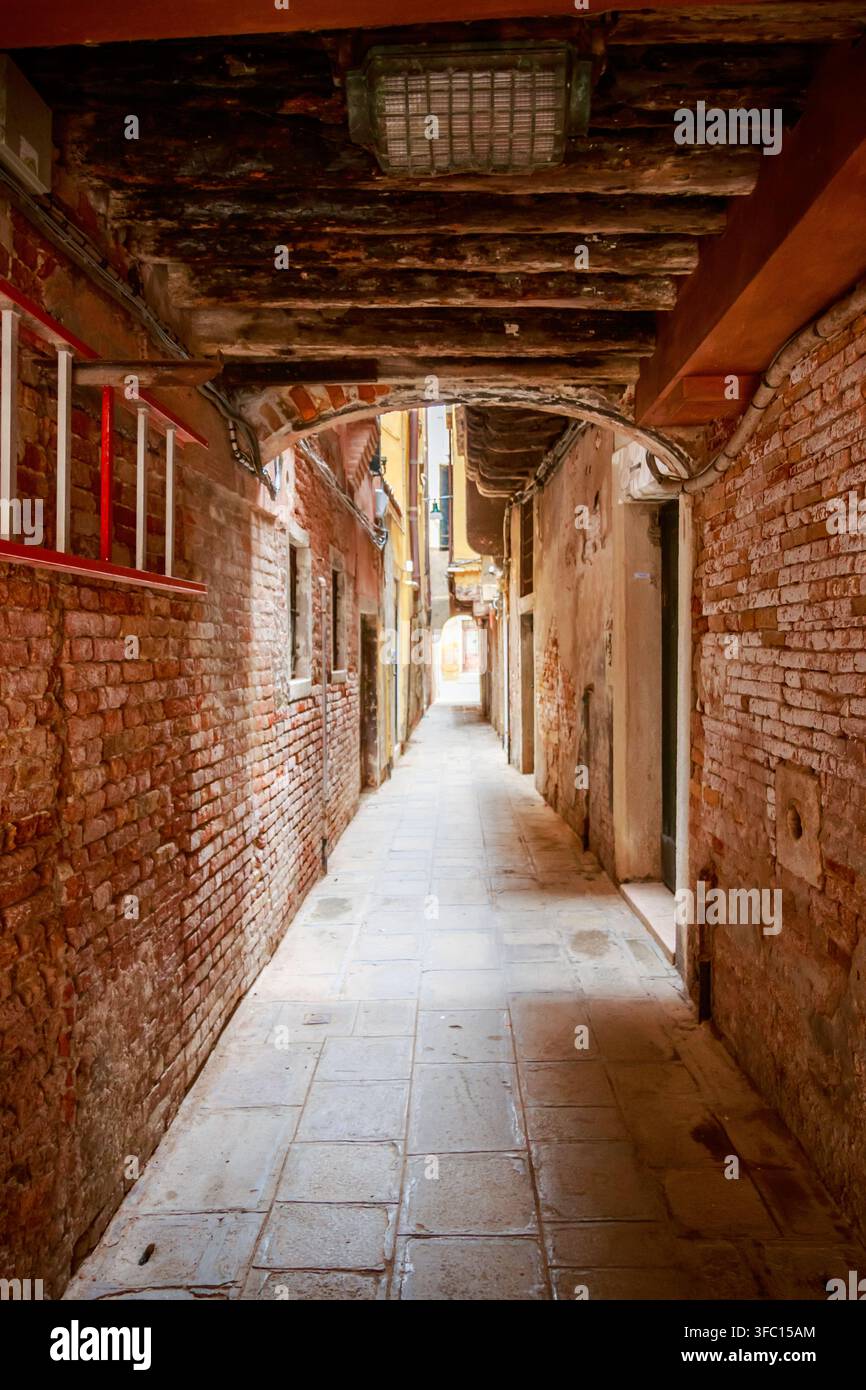Vicolo stretto di mattoni sotto vecchie travi di legno e un passaggio ad arco, con muri consumati, pavimentazione piastrellata e luce soffusa all'estremità. Foto Stock