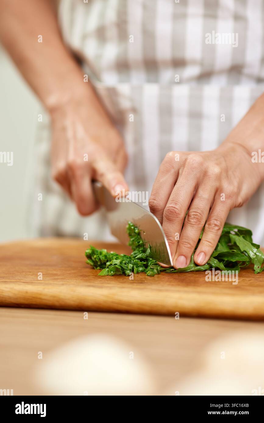 Mani che tengono un coltello grande, affettando la rucola verde fresca in piccoli pezzi su un tagliere di legno, preparando il cibo Foto Stock