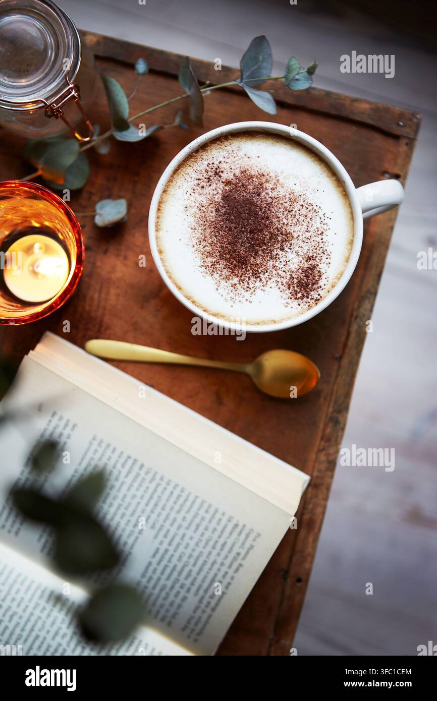 Una tazza di caffè ricoperta da una schiuma di latte cremosa e spolverata di cacao si trova su un tavolo di legno scuro. Accanto alla tazza ci sono un libro aperto e una candela accesa. Foto Stock