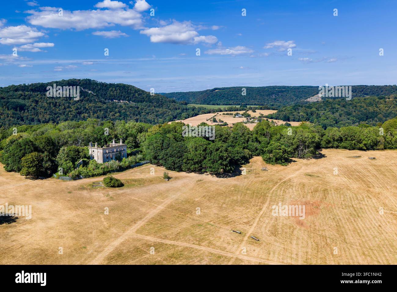 Rovine di una casa di campagna abbandonata del XVIII secolo incastonate tra alberi maturi, campi gialli aridi in primo piano e creste boschive sotto un cielo blu, Chepstow Foto Stock
