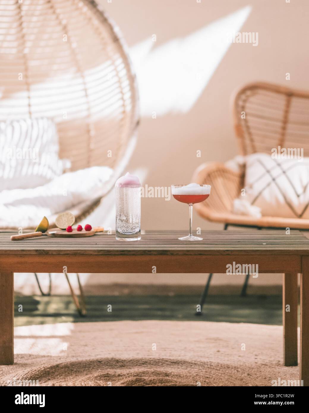 Cocktail coupe rosa e un bicchiere alto su un tavolo di legno tra sedie di rattan su una terrazza boho soleggiata, aperitivo estivo outdoor lifestyle Foto Stock