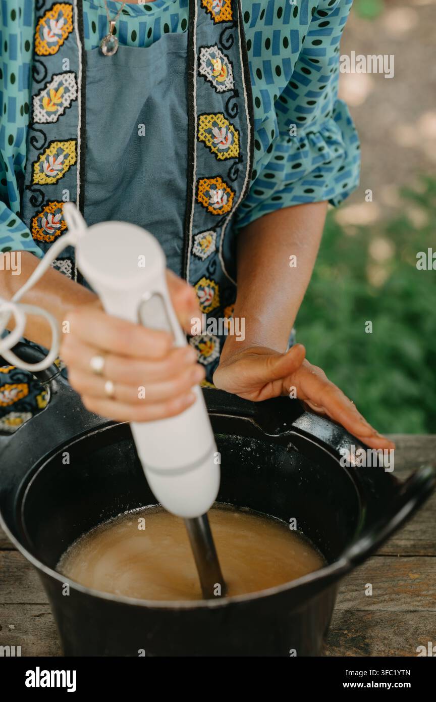 Prima piano di una persona che mescola una preparazione con un frullatore a mano in una pentola nera all'aperto accanto a un tavolo di legno rustico Foto Stock