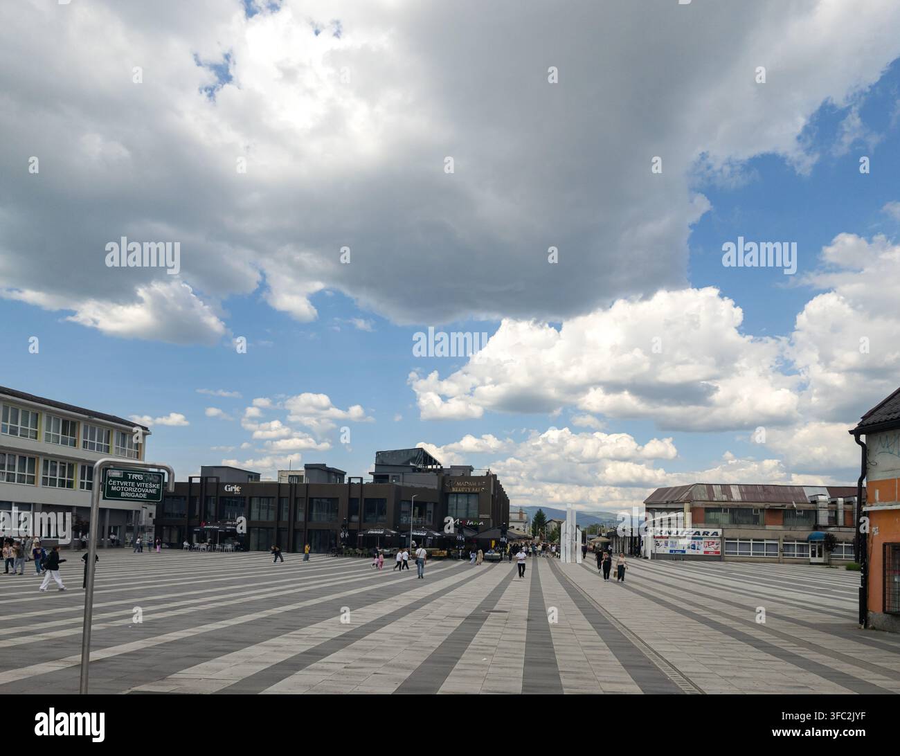 I pedoni attraversano una larga piazza moderna lastricata a Ilidza, in Bosnia, sotto un cielo brillante con drammatiche nuvole cumulus. - Immagine stock catturata con smartphone