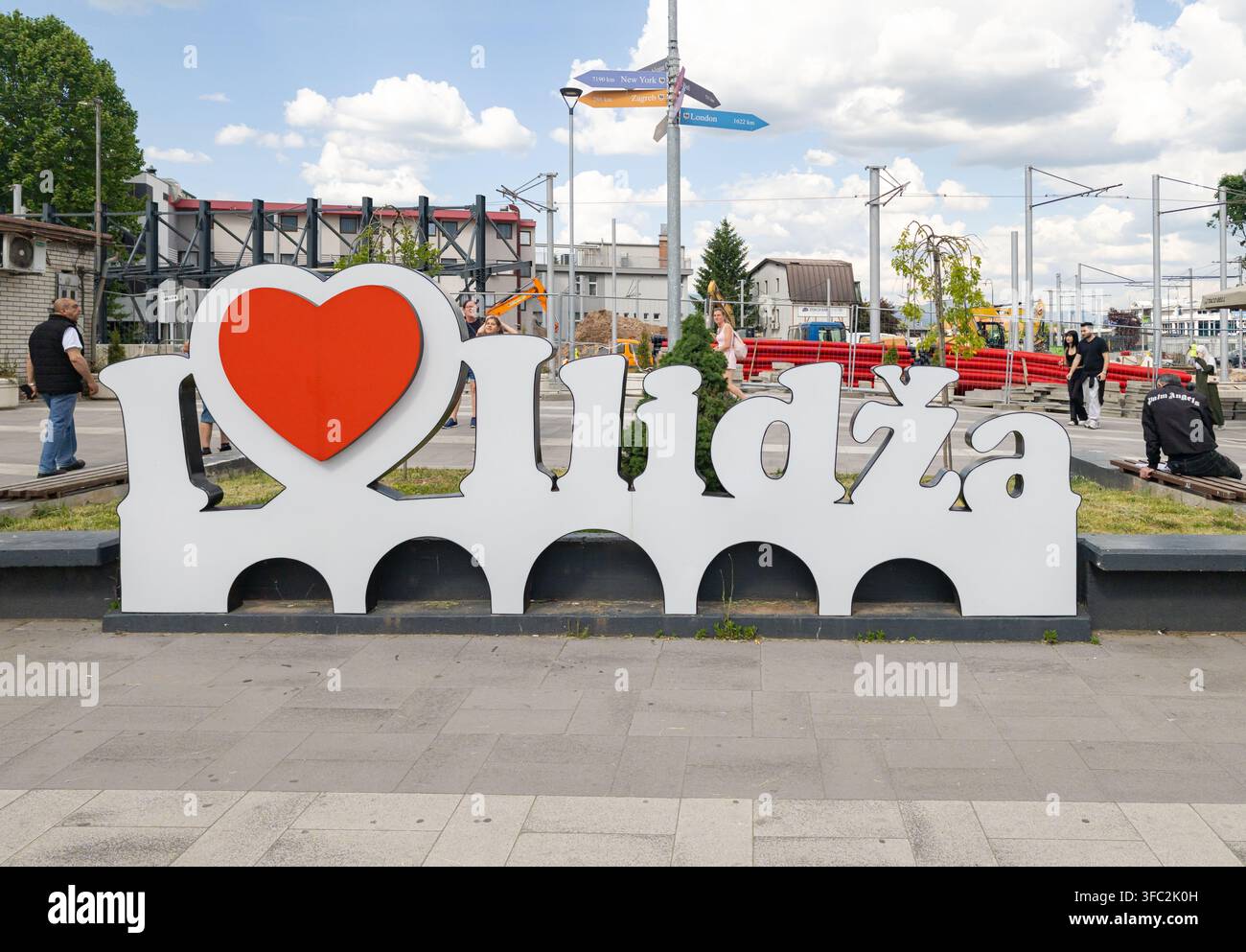 Un grande cartello bianco con la scritta "I Love Ilidza" si trova in una piazza pubblica a Ilidza, circondato da pedoni e segnali di distanza urbani. - Immagine stock catturata con smartphone