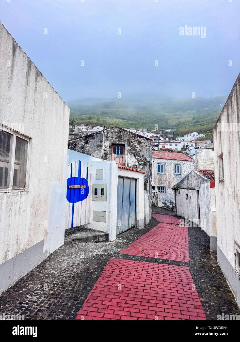 Corvo, Azzorre, Portogallo Un vicolo stretto a Corvo, Portogallo, presenta un sentiero in mattoni rossi. Muri imbiancati a calce delimitano entrambi i lati con porte blu e bianche. Nebbia Foto Stock