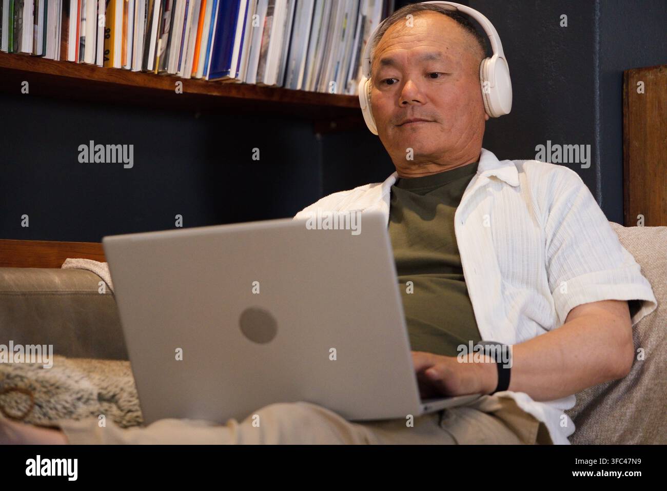 Uomo asiatico anziano seduto su un divano imbottito nel soggiorno utilizzando un laptop argentato e cuffie bianche Foto Stock
